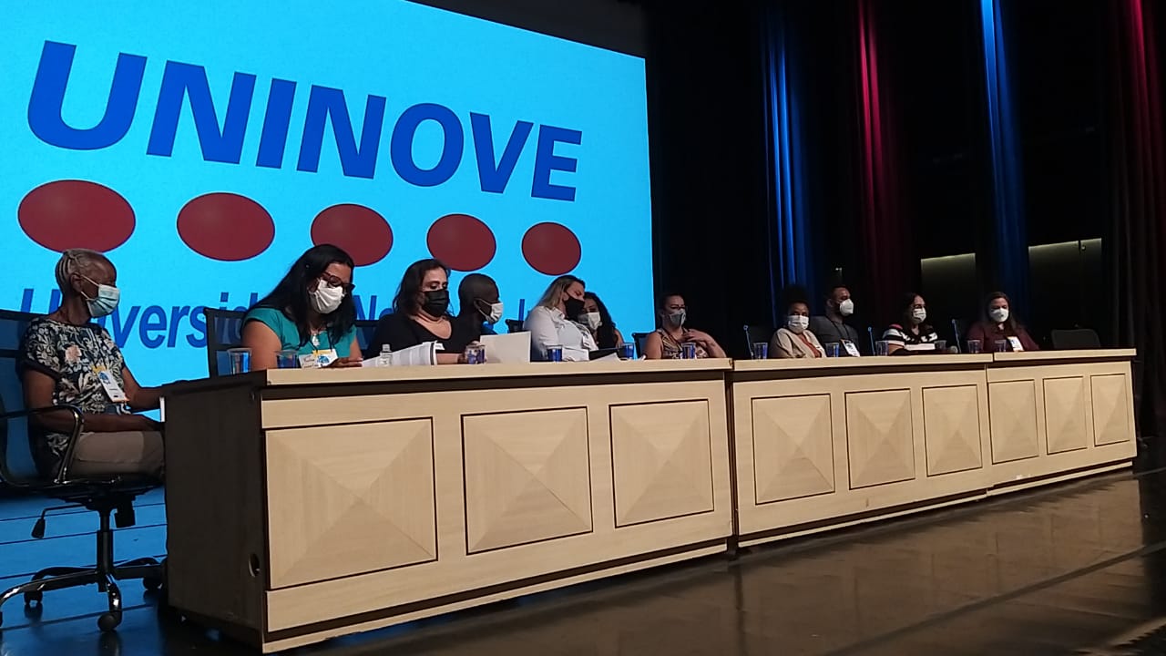 Em um palco há onze pessoas sentadas a mesa. Ao fundo tem projetor escrito Uninove.